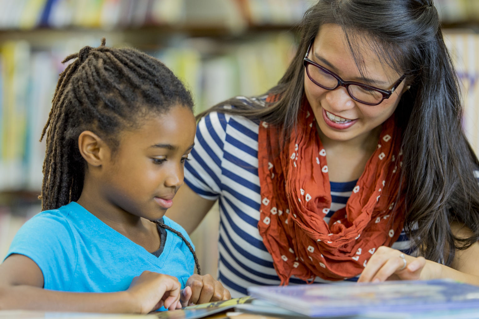 A teacher is reading books with an elementary age girl in the library.